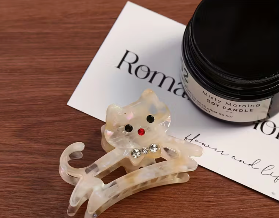 Small pearlescent acrylic cat hair clip with rhinestone bow displayed beside a soy candle on a wooden surface