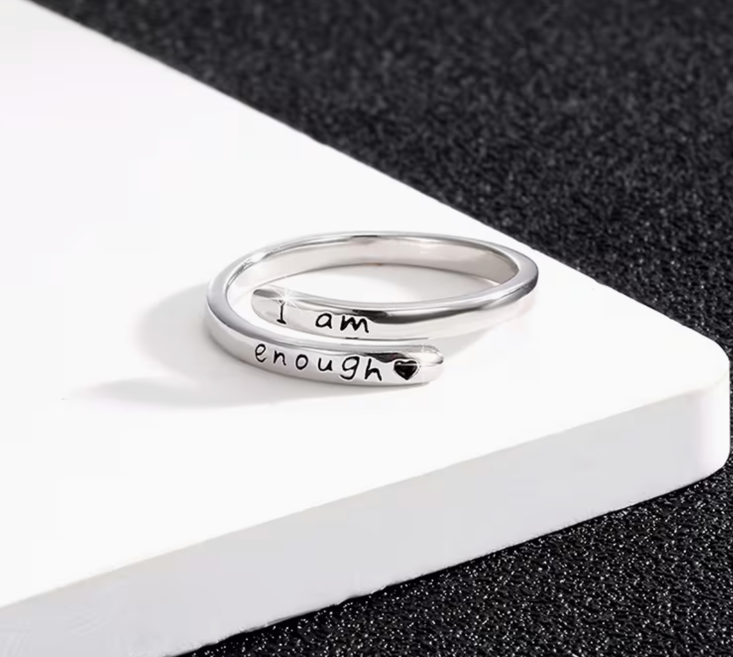 Adjustable silver tone affirmation ring engraved with the message I am enough displayed on a white platform against a dark textured background