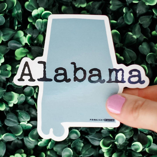 Alabama state outline vinyl sticker with waterproof and weatherproof finish held over green leafy background