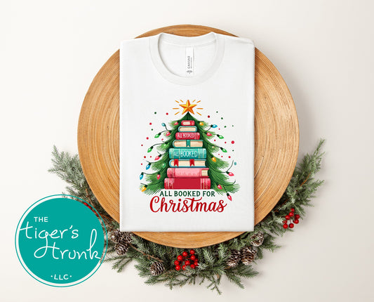 White short-sleeve shirt featuring a colorful Christmas tree made of stacked books with the phrase "All Booked for Christmas."