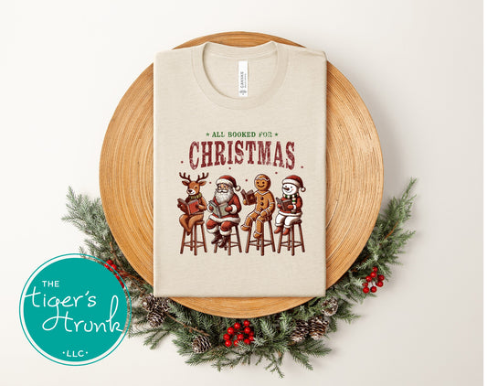 Natural heather short-sleeve shirt featuring Santa, a reindeer, gingerbread cookie, and snowman reading books beneath the phrase "All Booked for Christmas."
