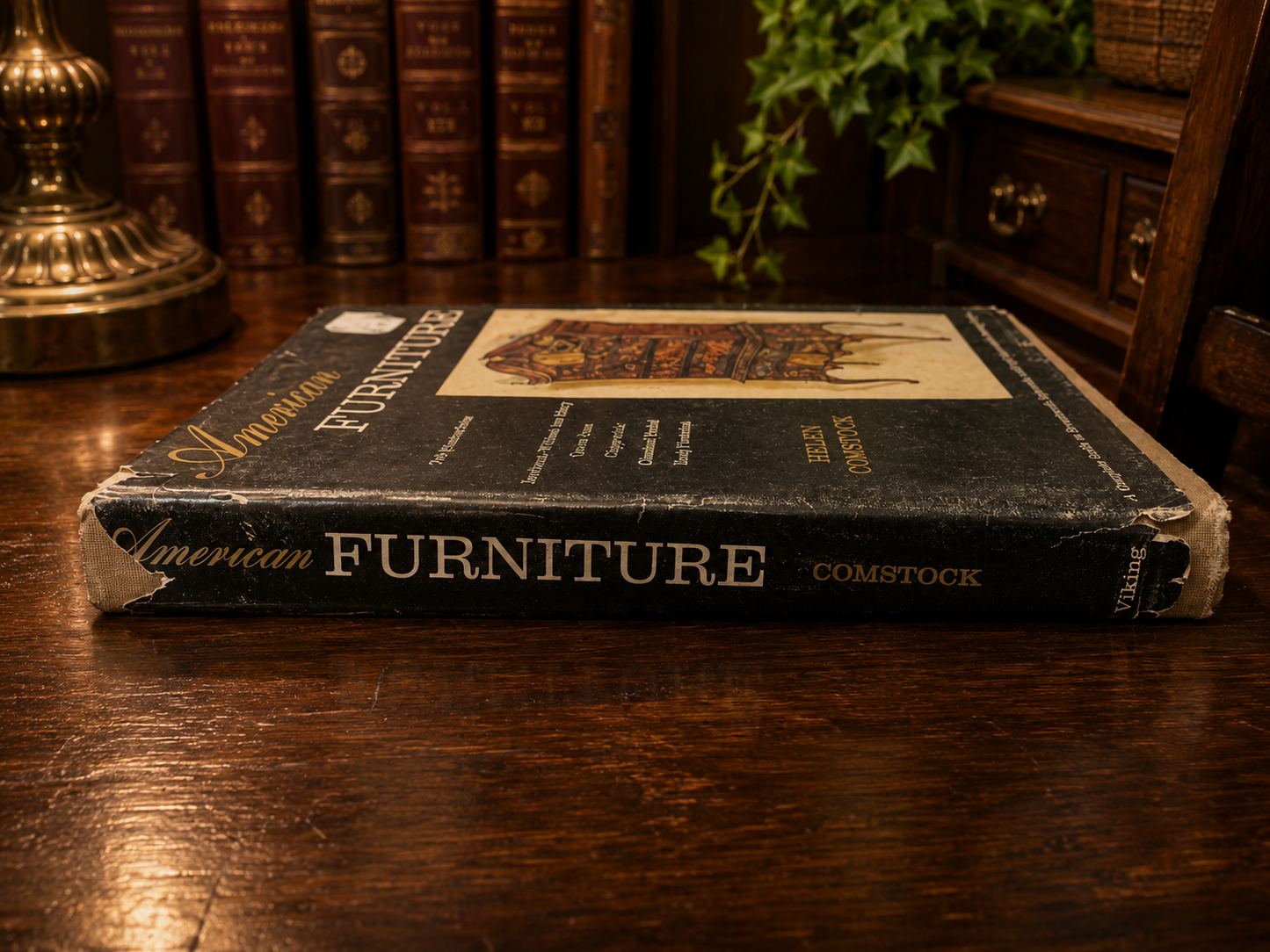 Book titled 'American Furniture' by Comstock on a wooden surface with books and a plant in the background.