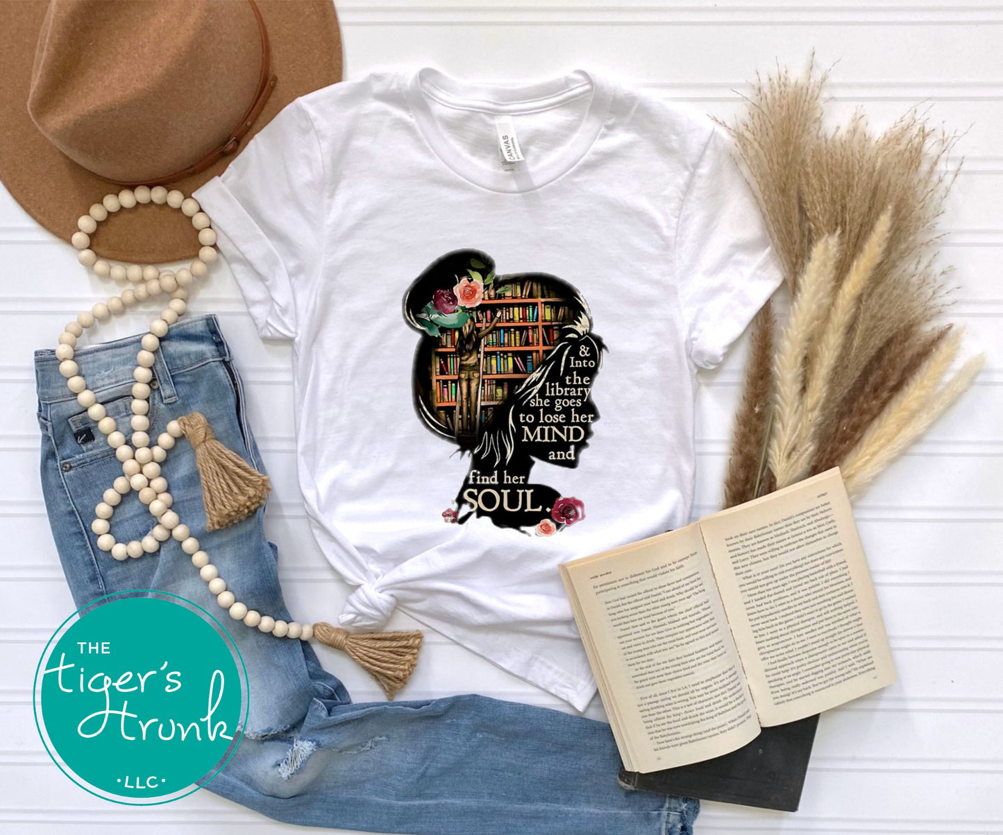 White t-shirt with a graphic design featuring a woman with books, surrounded by denim jeans, a hat, beads, and an open book on a white surface.