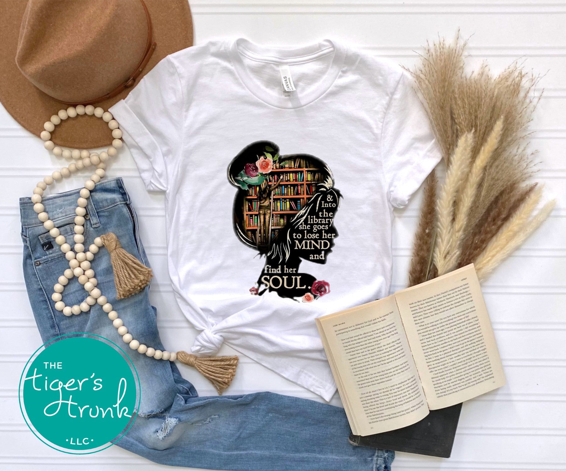White t-shirt with a graphic design featuring a woman with books, surrounded by denim jeans, a hat, beads, and an open book on a white surface.