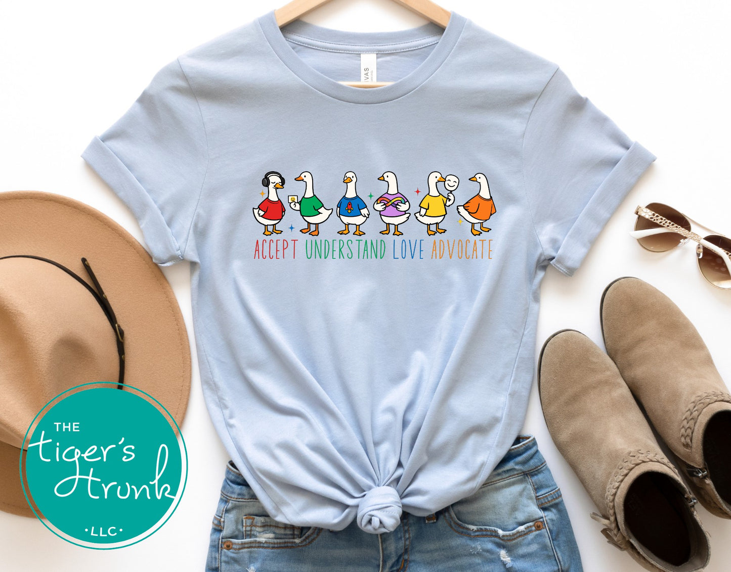 Light blue t-shirt with colorful duck graphic and text, displayed with a hat, sunglasses, and boots on a white background.
