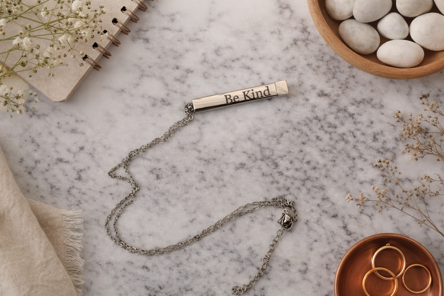Minimalist engraved bar necklace showing the phrase Be Kind on a sleek silver bar pendant resting on a marble background.
