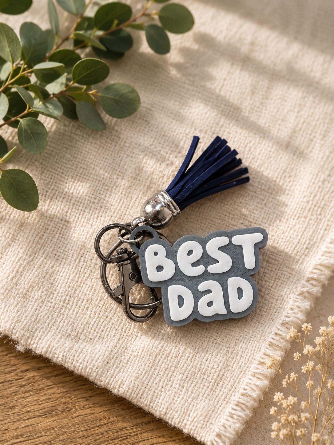 Gray and white Best Dad keychain with navy tassel displayed on textured neutral fabric