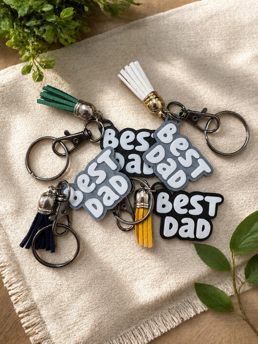 Collection of 3D printed Best Dad keychains with colorful tassels displayed on neutral fabric background