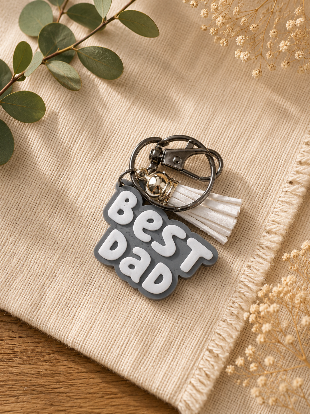Gray Best Dad keychain with white tassel displayed with eucalyptus and dried flowers on neutral cloth backdrop
