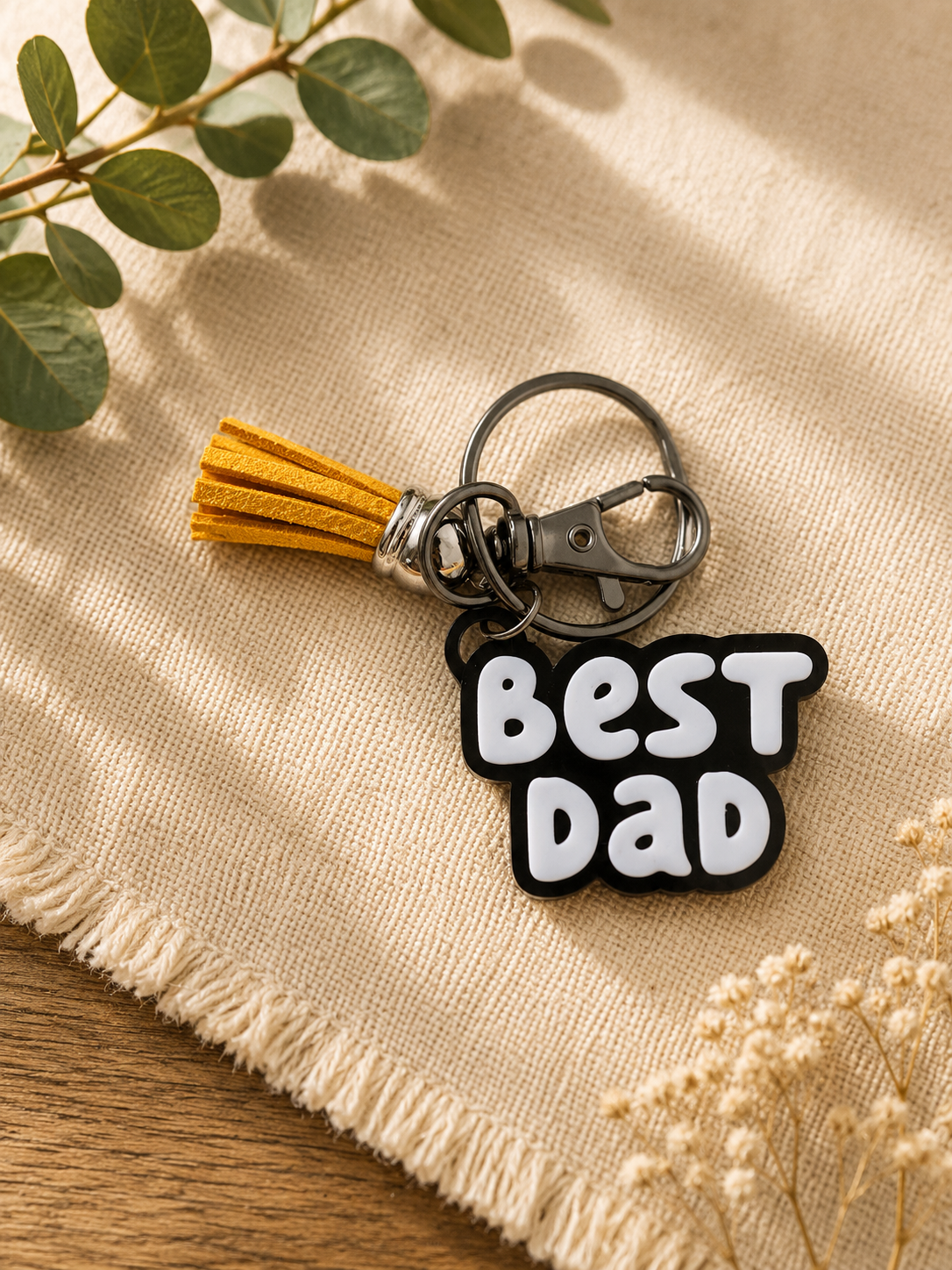 Black Best Dad keychain with yellow tassel and metal clip attachment on cream fabric background