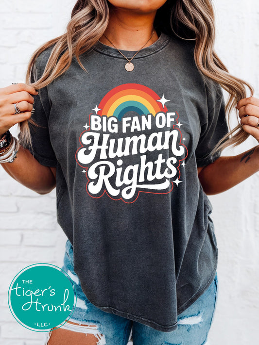Person wearing a t-shirt with 'Big Fan of Human Rights' design and rainbow, against a white brick wall.