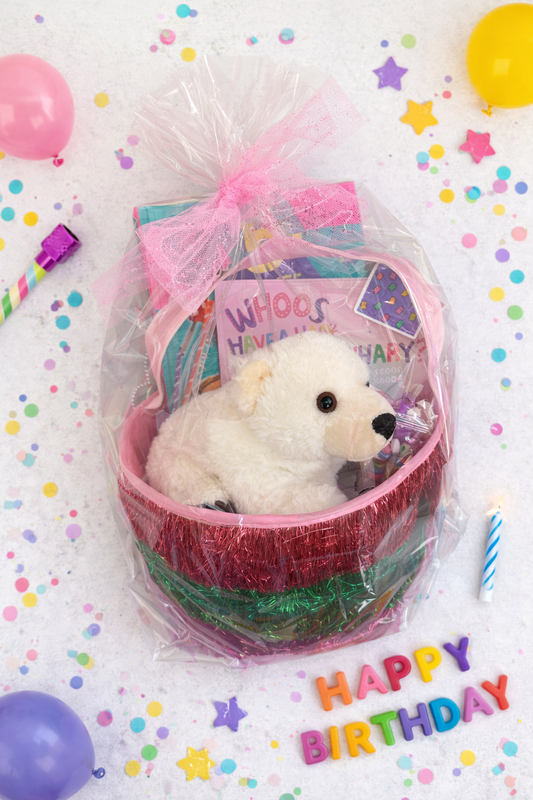 Pink birthday gift basket wrapped in clear cellophane with a plush white bear, children’s books, candy treats, and party decorations including confetti, balloons, and a lit candle.