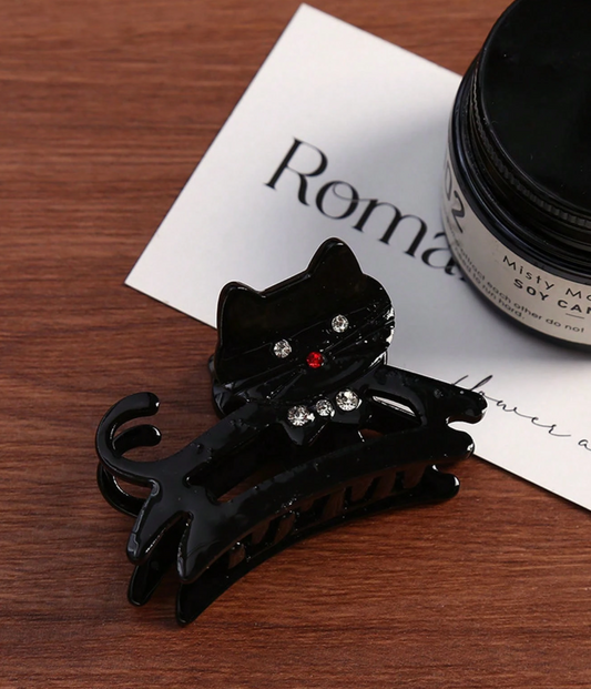 Black cat-shaped hair clip on a wooden surface with a white card and black container in the background.