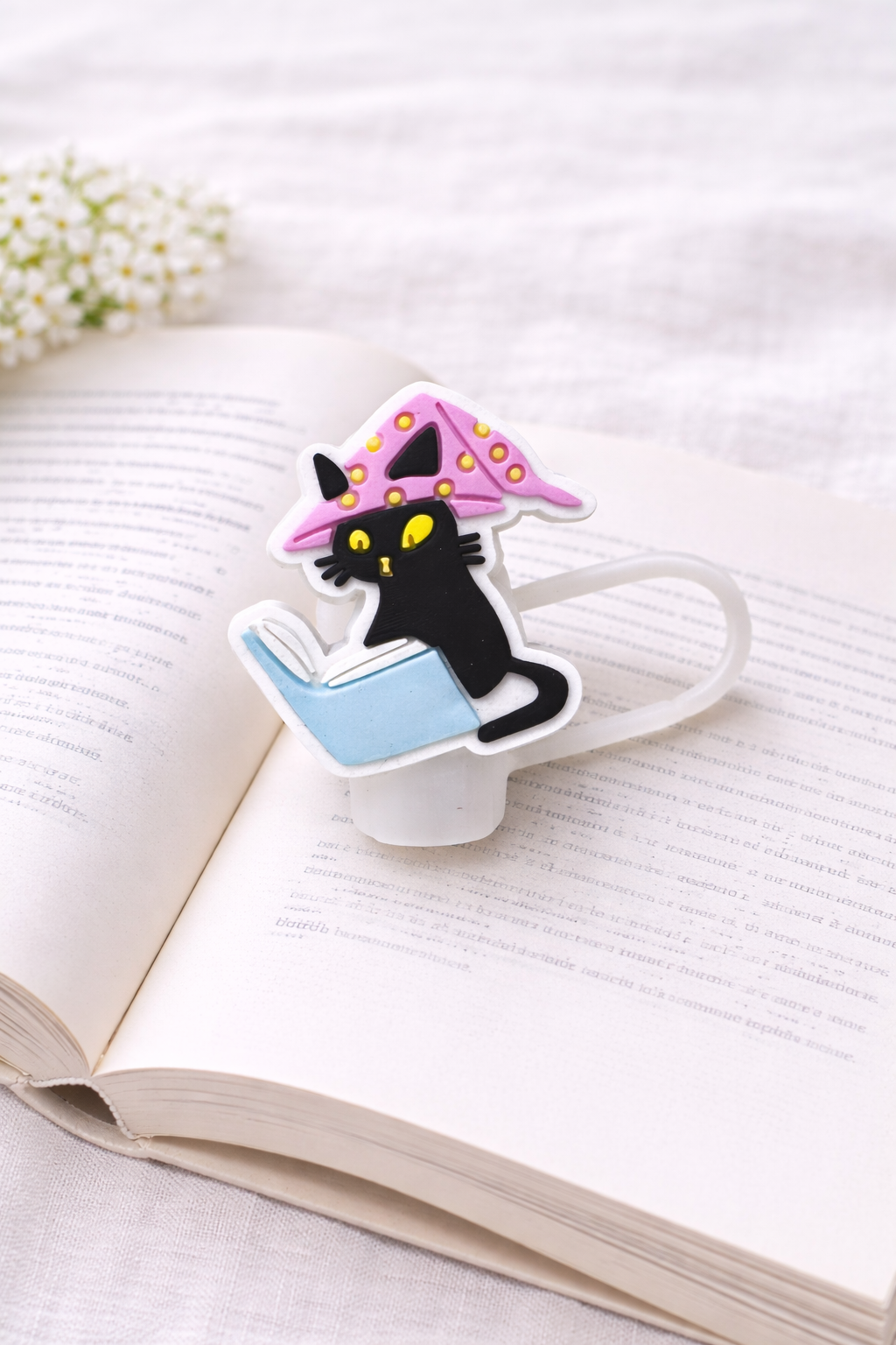 Black cat silicone straw topper wearing a pink hat while reading a blue book, displayed on an open book with soft floral background.