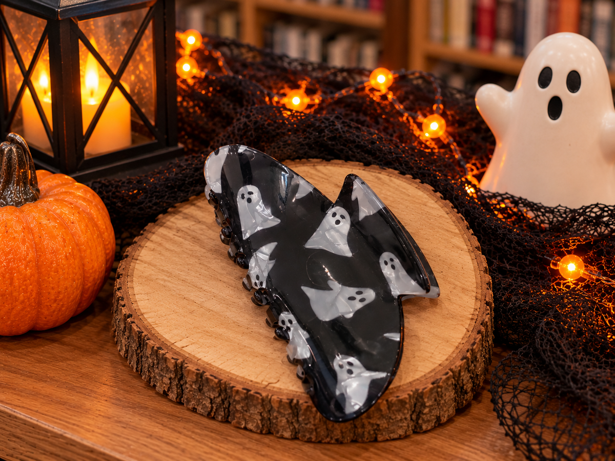 Black Halloween hair claw clip with white ghost designs, glossy marbled finish, and curved claw shape displayed on a wooden stand.