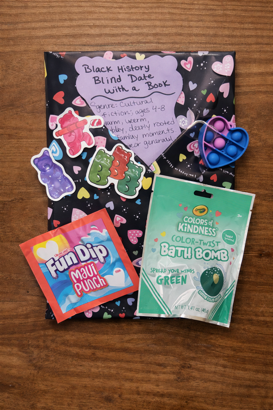 Flatlay of a Black History Blind Date with a Book bundle for ages 4–8 wrapped in heart-pattern paper, featuring gummy bear stickers, a heart-shaped fidget pop toy, green bath bomb, and Maui Punch Fun Dip on a wooden background.