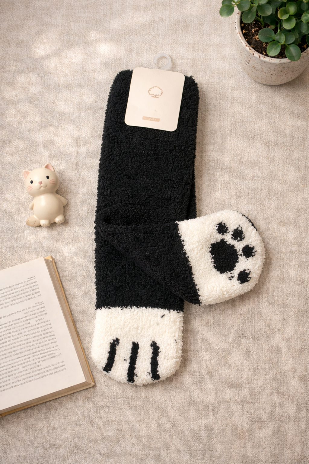 Flat lay of black and white fuzzy cat paw socks with paw pad design and claw toe markings, styled with a small cat figurine, open book, and plant on a neutral fabric background.
