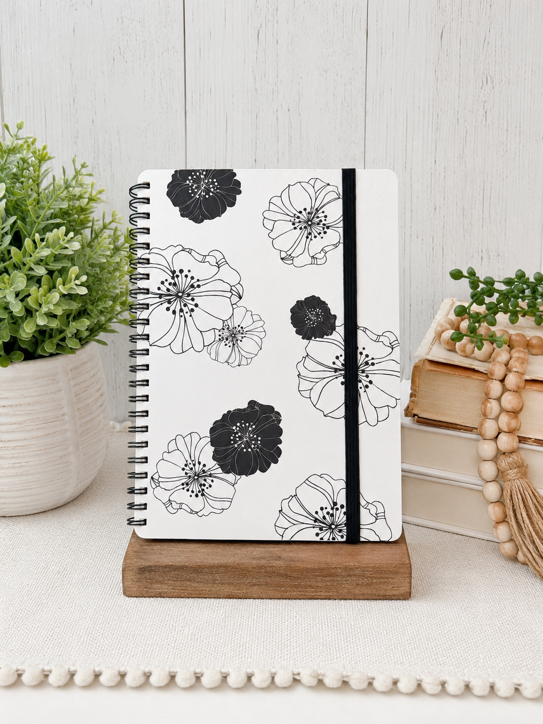 Black and white floral spiral journal with elastic closure and lined pages displayed on a wooden stand.