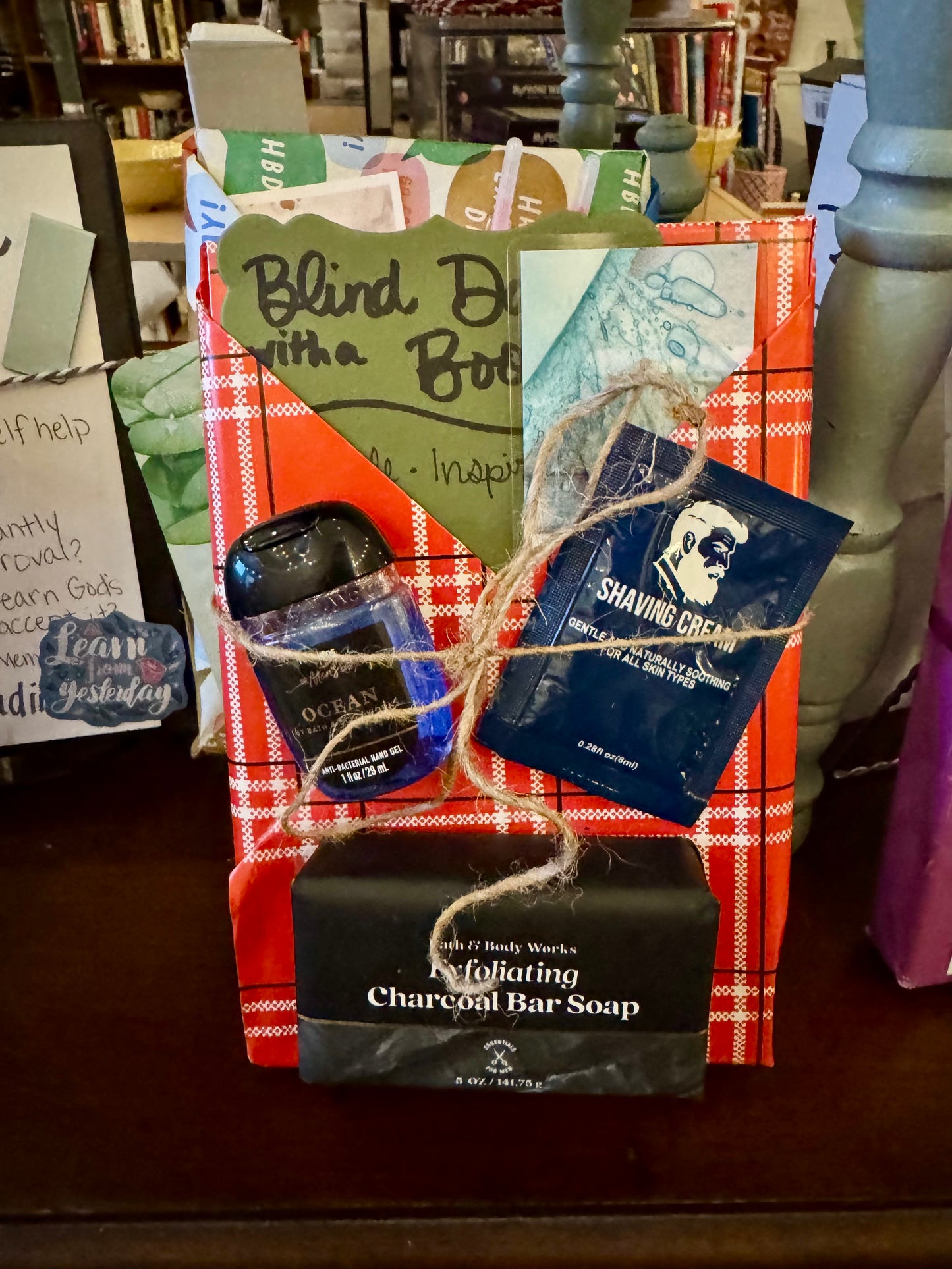 Front view of a Blind Date with a Book gift wrapped in red plaid paper, featuring a parable-inspired book bundle with exfoliating charcoal bar soap, shaving cream sample, and hand sanitizer tied with twine, displayed inside an independent bookstore.