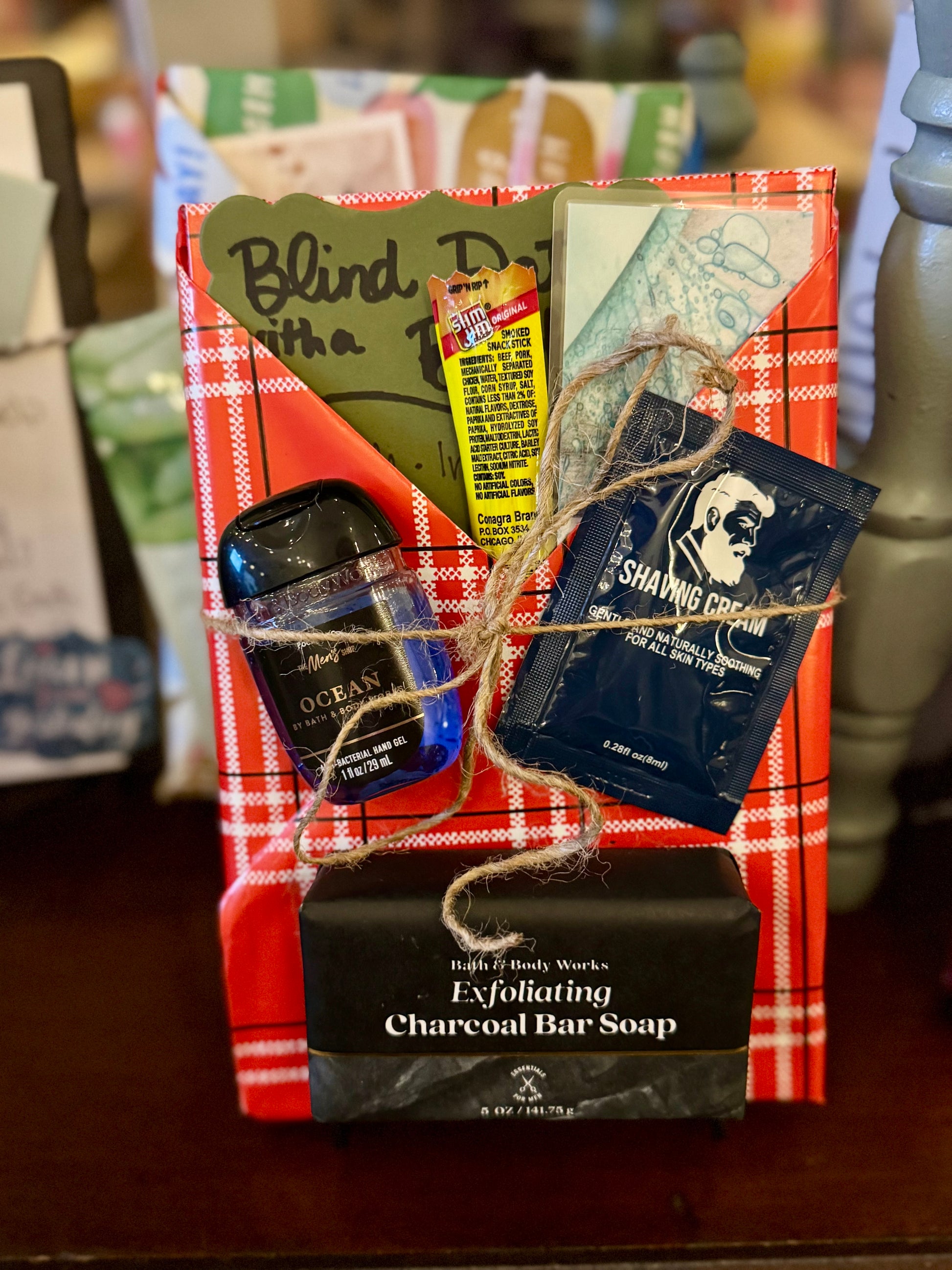 Blind Date with a Book gift wrapped in red plaid paper featuring a parable-inspired inspirational fiction book, exfoliating charcoal bar soap, shaving cream sample, hand sanitizer, and bookmark tied with twine, displayed on a wooden surface inside a bookstore.