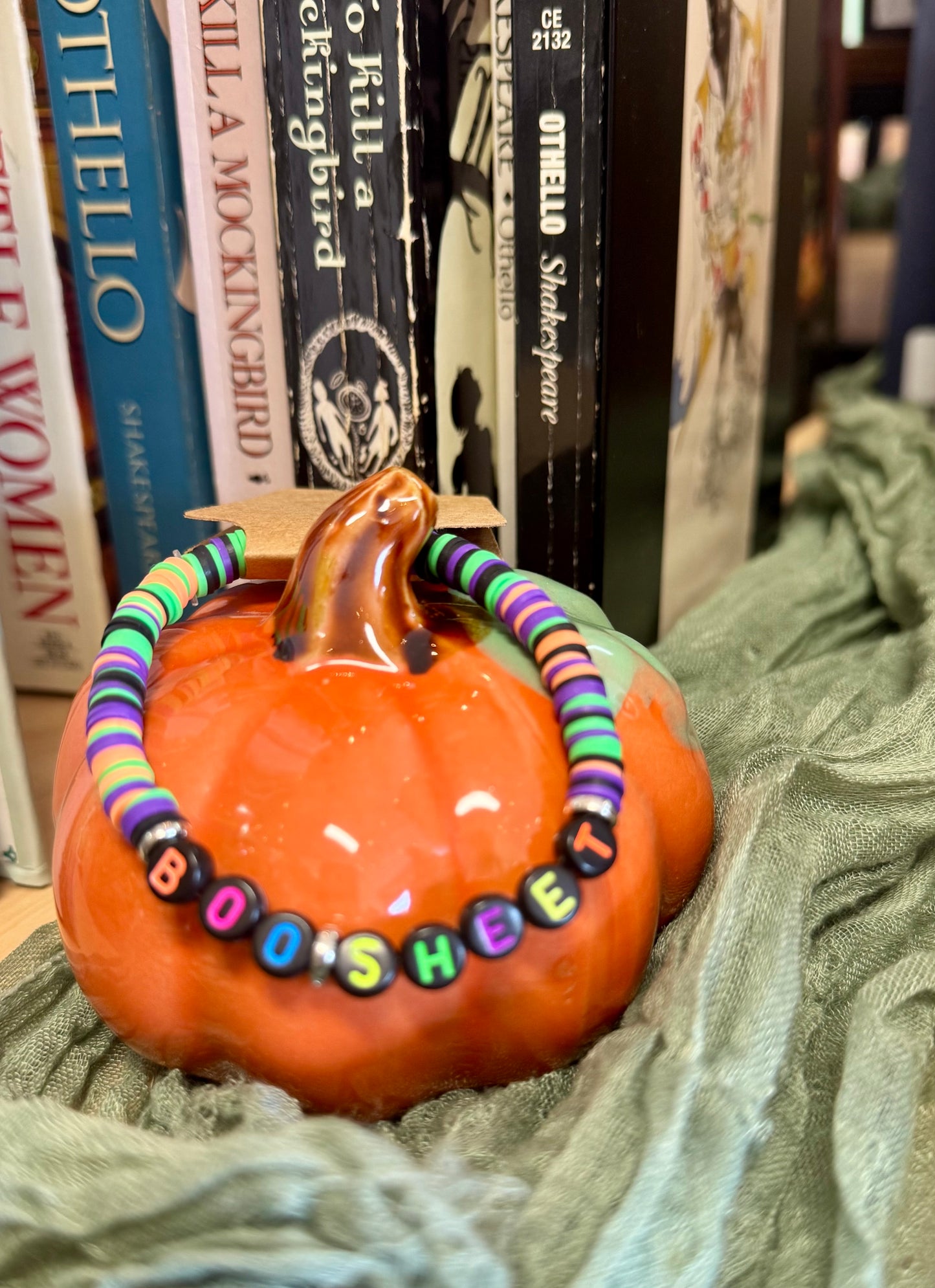 Pumpkin with a colorful 'BOO SHEE' beaded necklace on a green surface with books in the background.