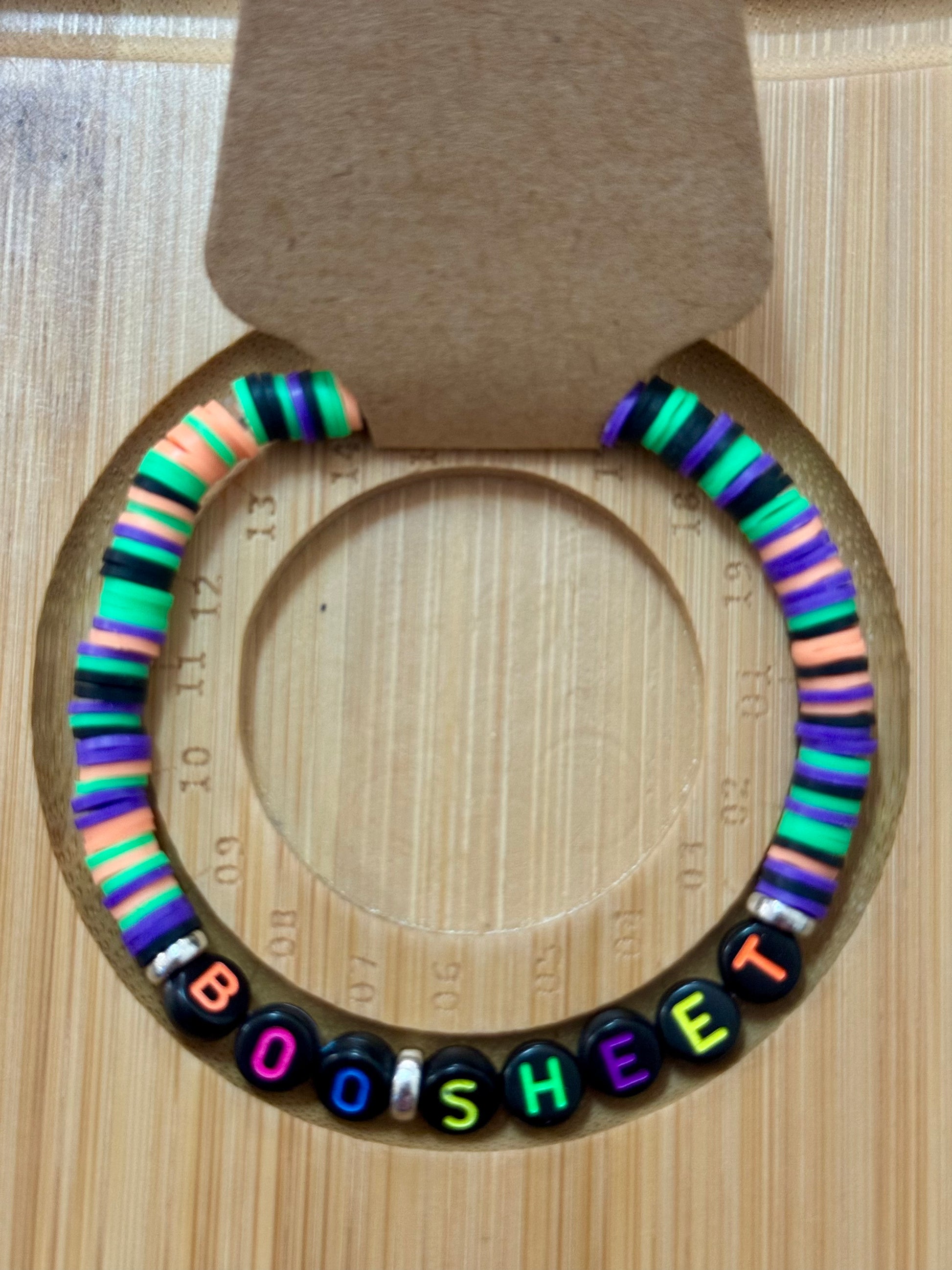 Colorful beaded bracelet with letter beads spelling 'BOO SHEET' on a wooden surface.
