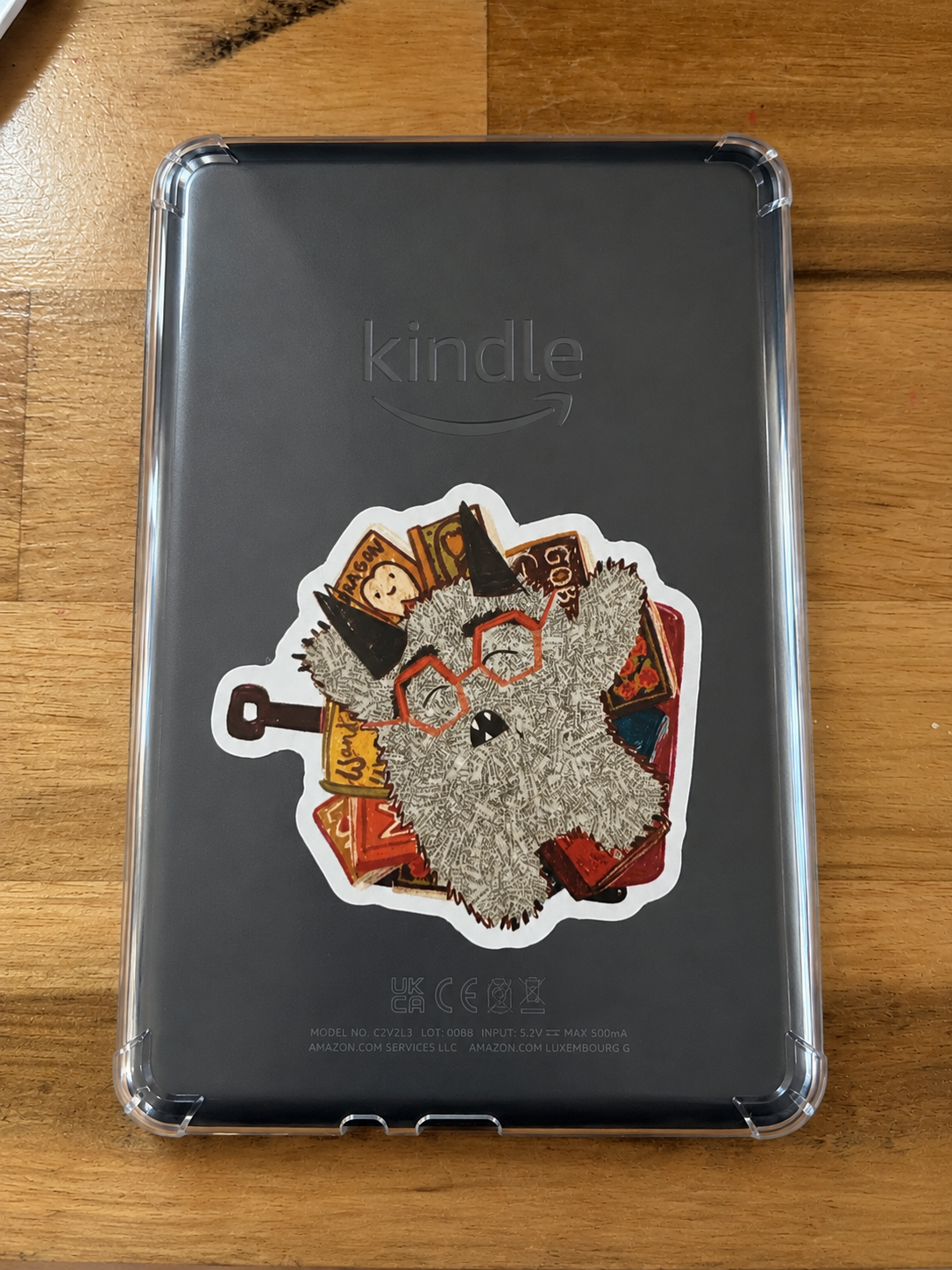 Cute fuzzy Book Monster character surrounded by books displayed as a vinyl sticker on the back of a Kindle inside a clear case.
