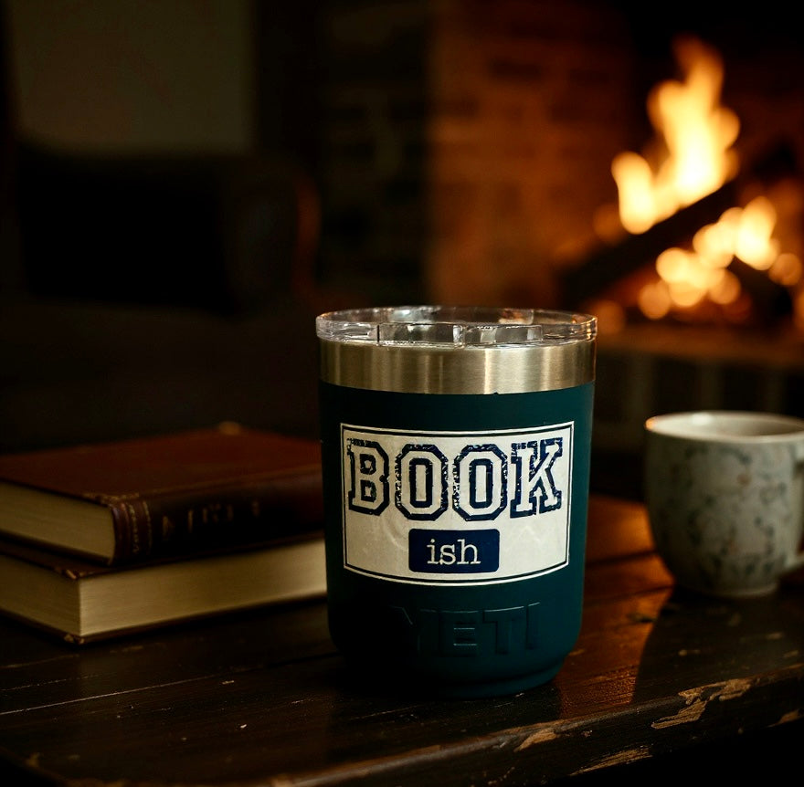 dark blue insulated tumbler with bookish design on a wooden table beside books and a fireplace