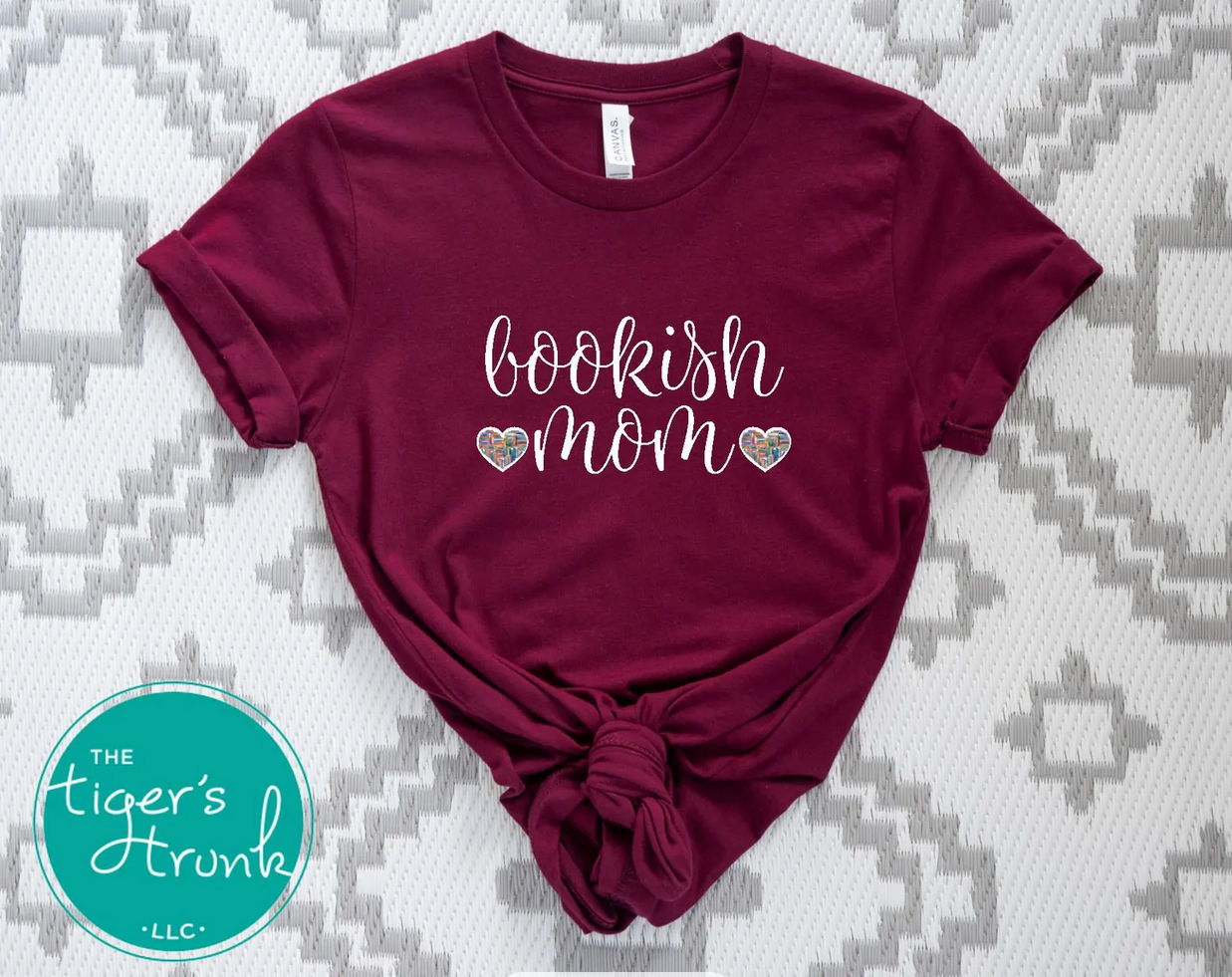 Maroon t-shirt with 'bookish mom' text on a patterned surface, featuring The Tiger's Trunk logo.