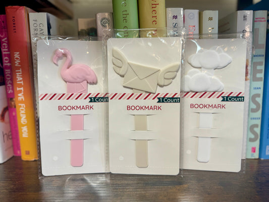 Three bookmarks in packaging with a pink flamingo, beige envelope, and white sheep design on a bookshelf background.