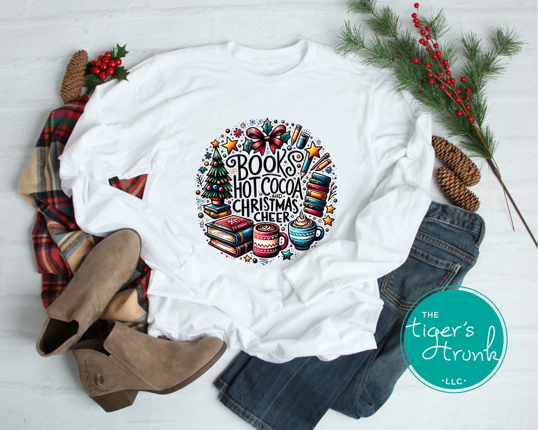 White long-sleeve shirt featuring Books Hot Cocoa and Christmas Cheer graphic, paired with jeans, boots, plaid blanket, and holiday greenery.