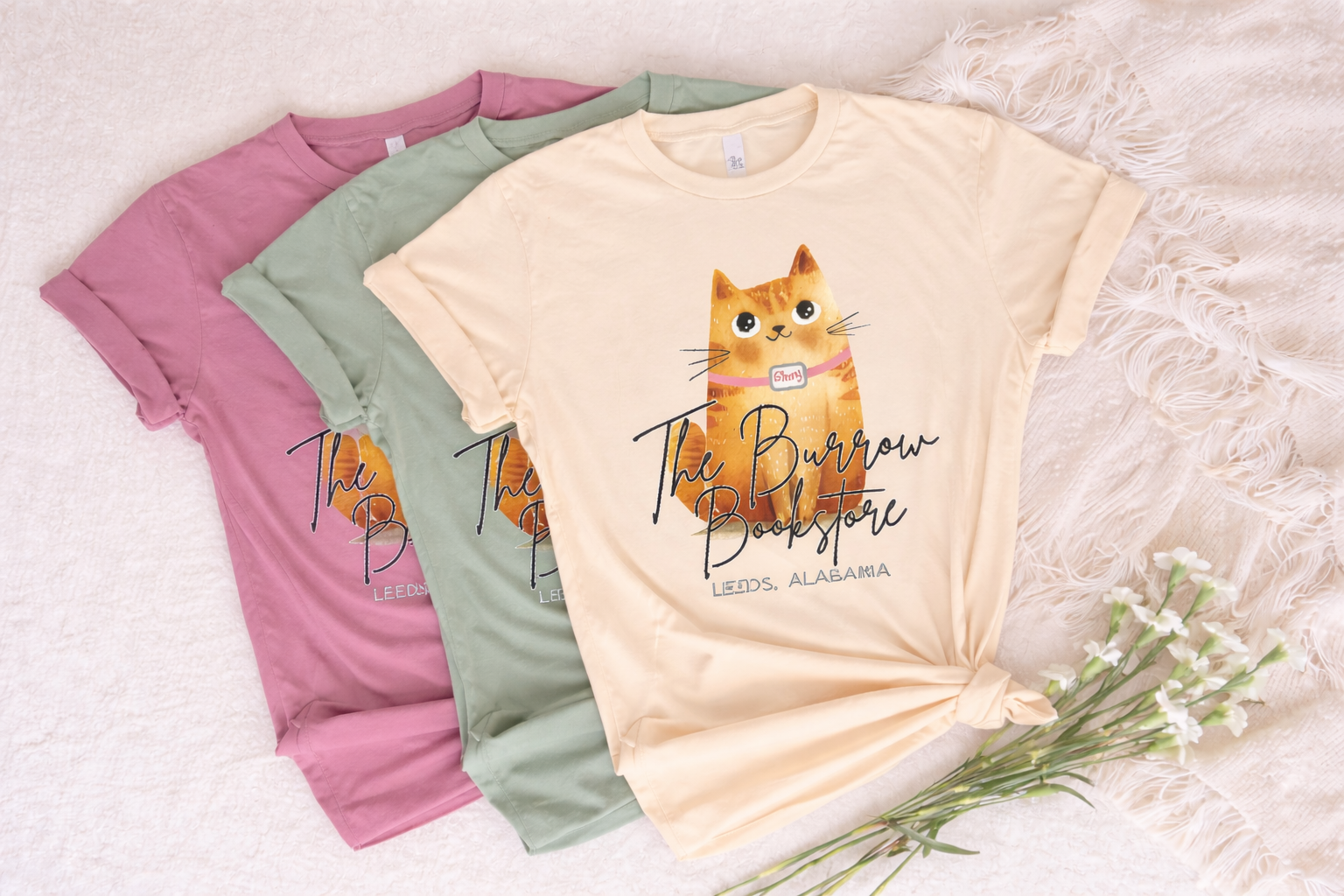 Three t-shirts with a cat graphic and text on a textured surface with flowers.