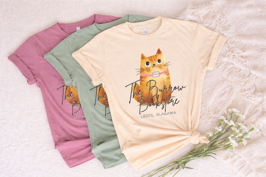Three t-shirts with a cat graphic and text on a textured surface with flowers.