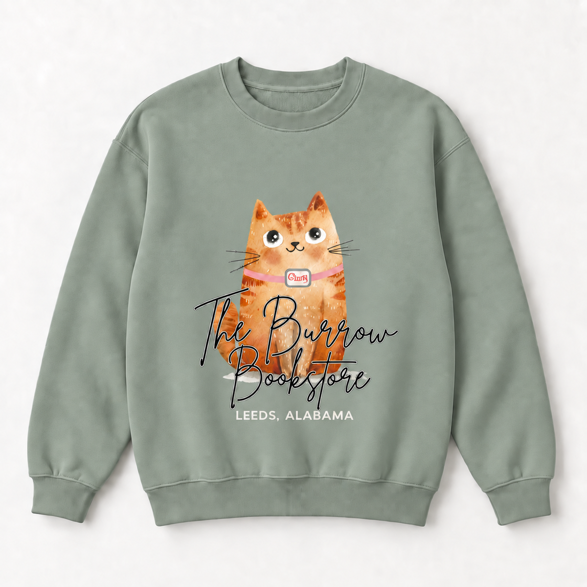A sage green crewneck sweatshirt featuring a watercolor-style orange tabby cat graphic with the text 'The Burrow Bookstore' and 'LEEDS, ALABAMA' printed on the front, displayed against a light gray background.