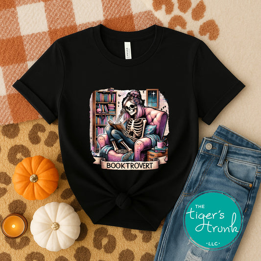 Black t-shirt with skeleton reading a book design, placed on a checkered surface with pumpkins and jeans.