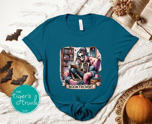 Blue t-shirt with a skeleton reading a book design, placed on a textured surface with decorative items.