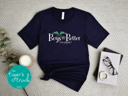 A maroon short-sleeve t-shirt with the phrase 'Boys are better (on paper)' printed on it, displayed on a flat surface with a book, glasses, and a plant.