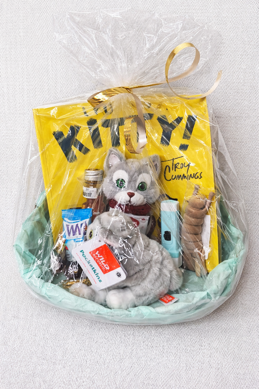 Cat-themed gift basket with plush gray kitty, snacks, cookies, and small self care items wrapped in clear gift wrap with ribbon