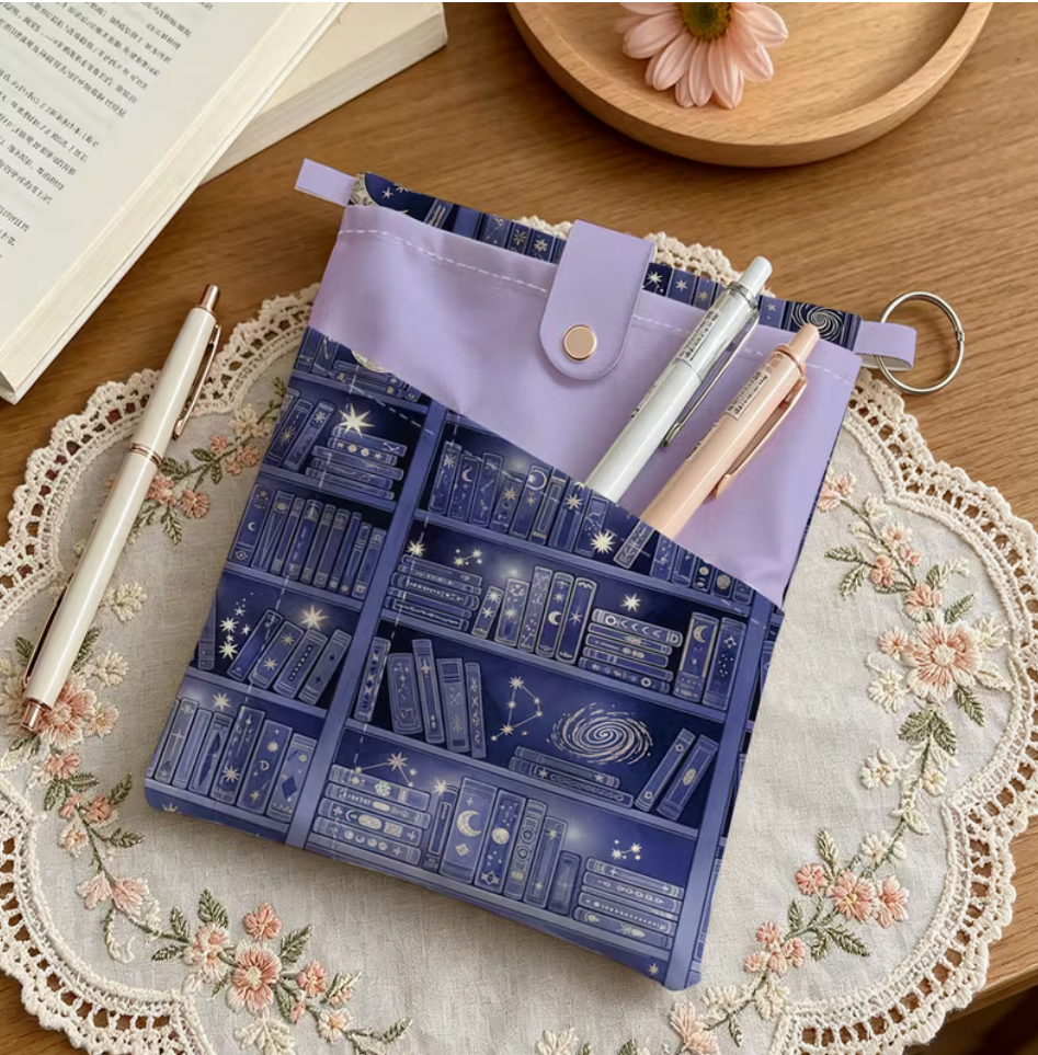 Navy celestial bookshelf book sleeve with lavender pen pockets, snap closure and pens displayed on a wooden table.