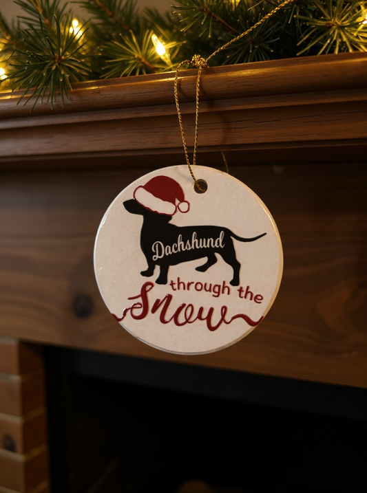 Ceramic Christmas ornament with a dachshund silhouette wearing a Santa hat and the phrase dachshund through the snow.