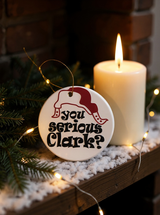 Ceramic Christmas ornament with a Santa hat graphic and the phrase you serious Clark, displayed near a candle and holiday greenery.
