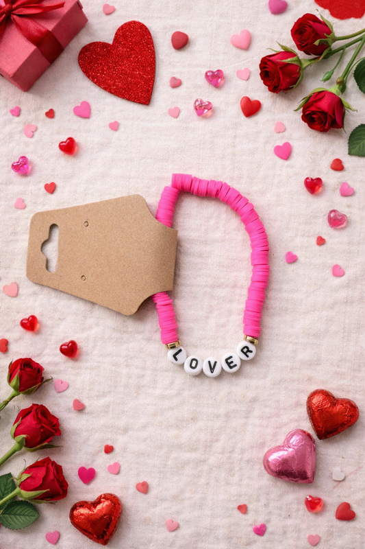 Pink clay beaded stretch bracelet with white letter beads spelling LOVER on a Valentine themed flat lay