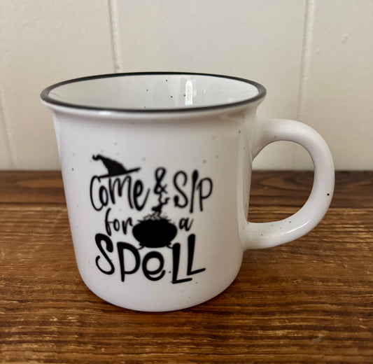 White mug with black text 'Come & Sip for a Spell' on a wooden surface.