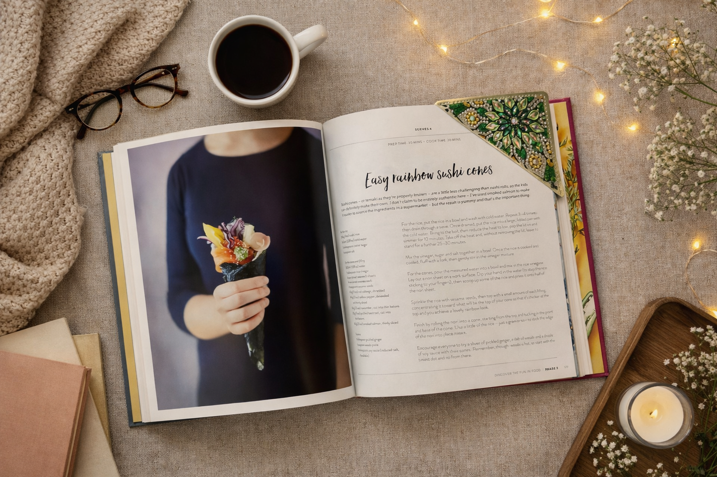 Overhead bookish flatlay of an open cookbook on neutral fabric, featuring a green jeweled corner bookmark clipped to the top right page, with a rainbow sushi cone recipe photo, coffee, glasses, and soft lights styled around the book.