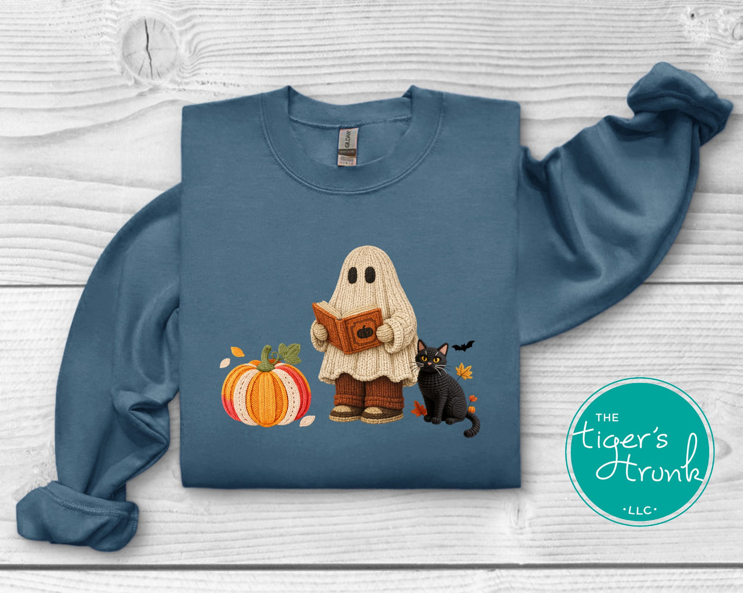 A folded slate blue sweatshirt on a white wood background, featuring a graphic of a ghost character reading a book next to a pumpkin and a black cat. A teal circular logo for 'The Tiger's Trunk LLC' is visible on the bottom right.