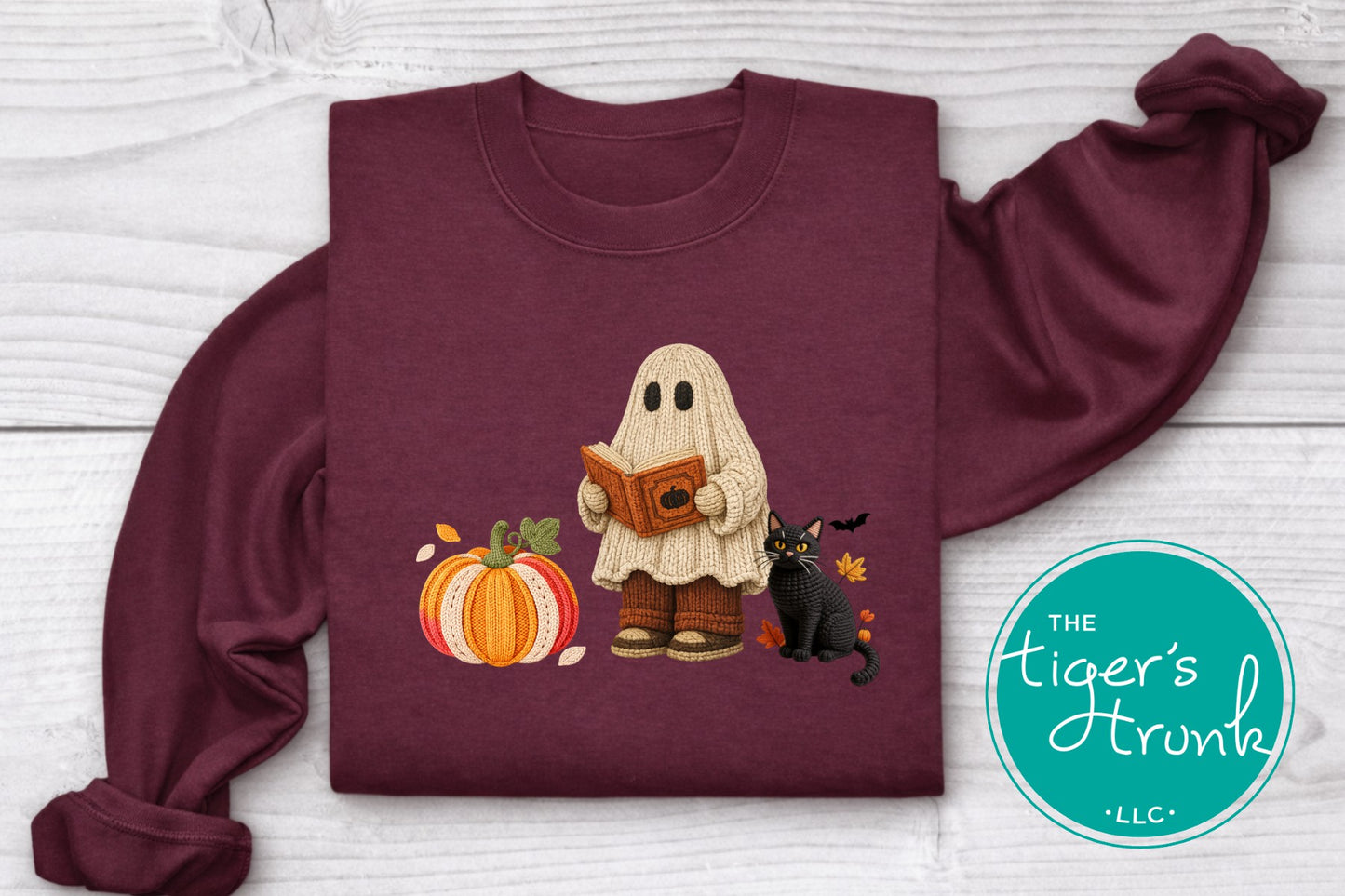 A folded maroon long-sleeve shirt with a graphic of a ghost reading a book next to a pumpkin and a black cat, displayed on a white wood background with a teal circular logo in the corner.