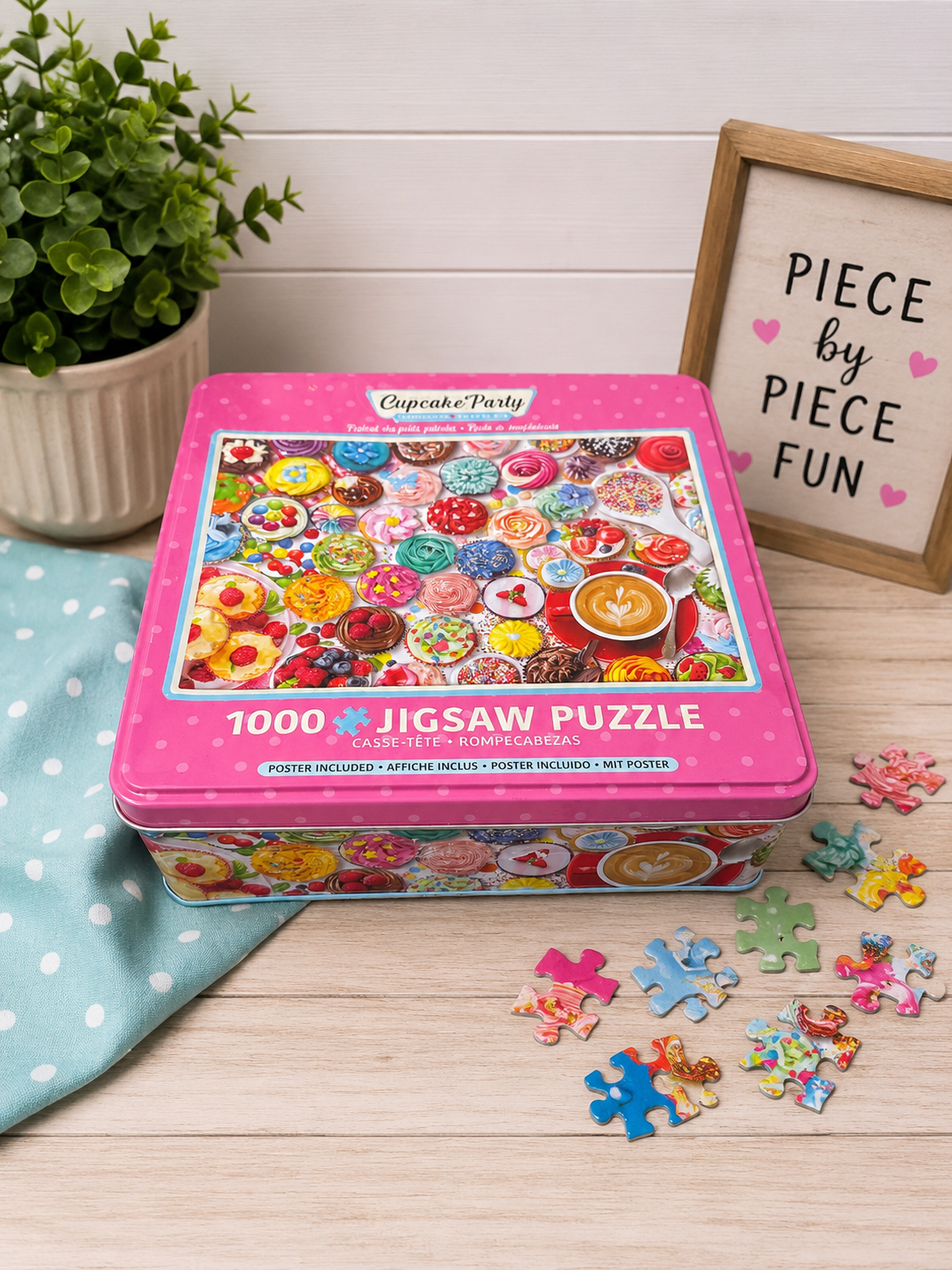 Cupcake Party 1000-piece jigsaw puzzle in a pink collectible tin displayed on a light wood surface with colorful puzzle pieces, a small potted plant, a soft blue polka-dot cloth, and a framed "Piece by Piece Fun" sign in the background.