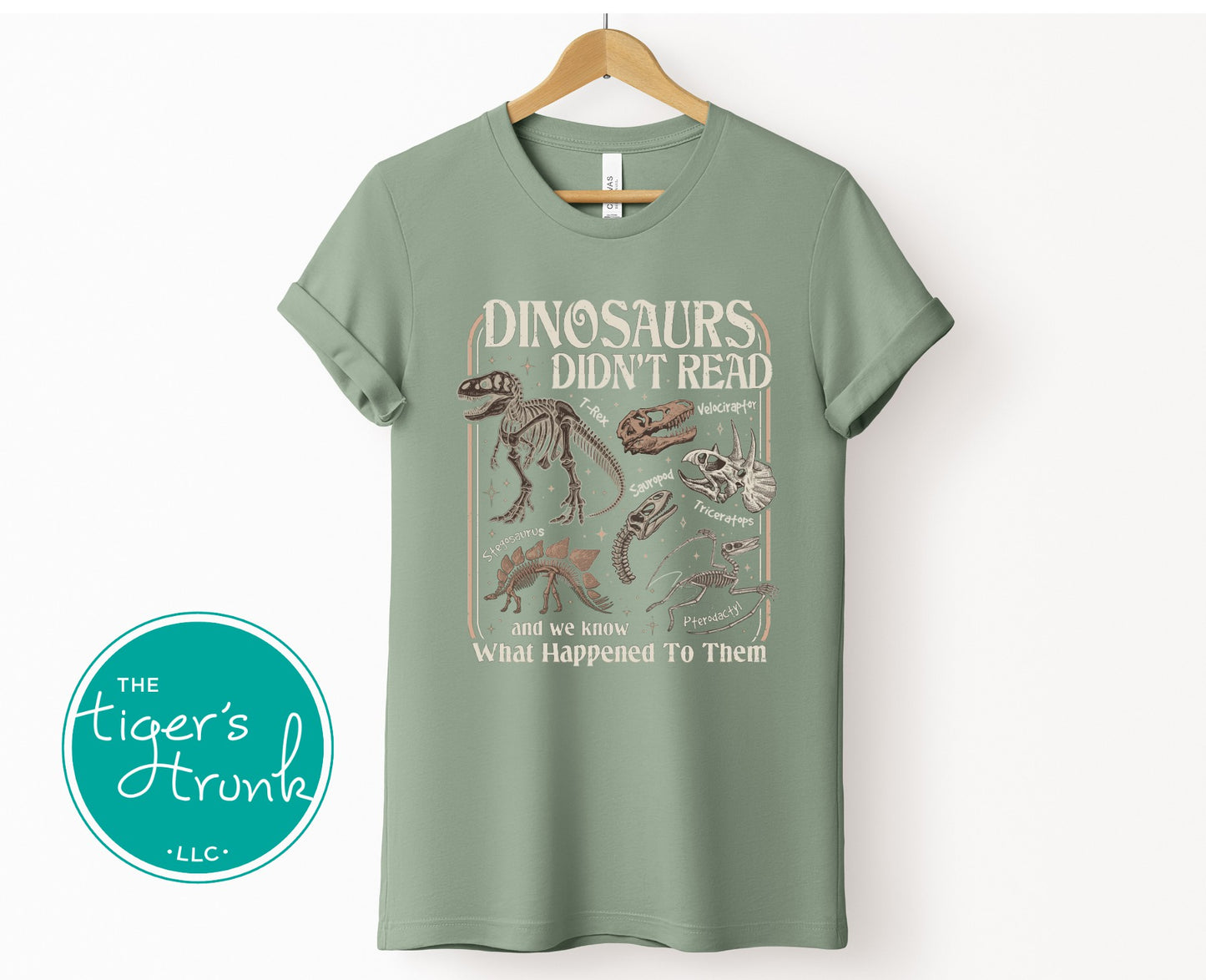 Sage green graphic t-shirt with dinosaur fossil illustrations and the phrase Dinosaurs Didn’t Read and We Know What Happened To Them