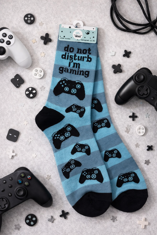Flat lay of blue Do Not Disturb I’m Gaming crew socks with black video game controller pattern, styled with game controllers and gaming accessories on a light background.