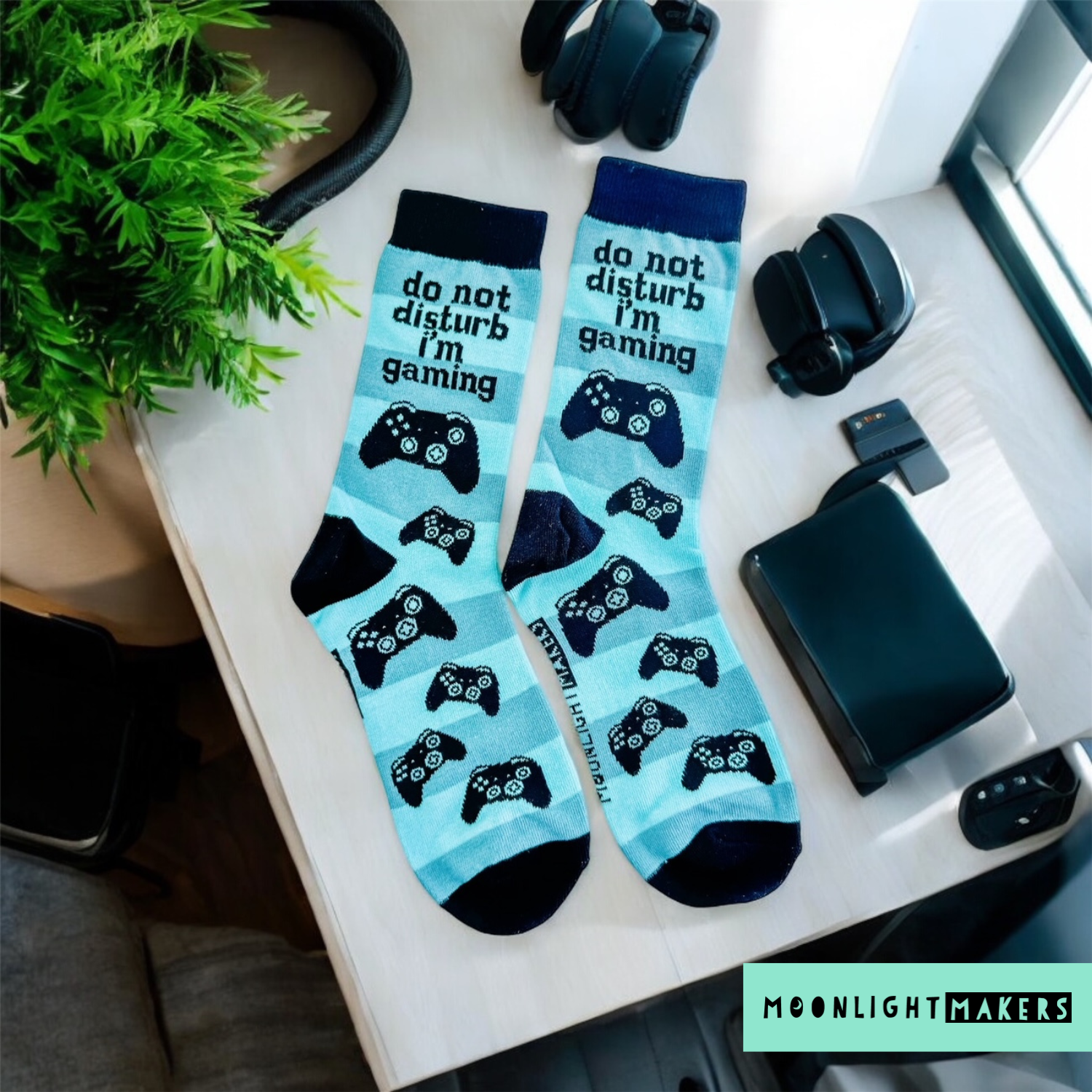 Do Not Disturb I’m Gaming light blue crew socks with black game controller pattern styled on a desk flatlay with headphones, phone, plant, and gaming accessories.